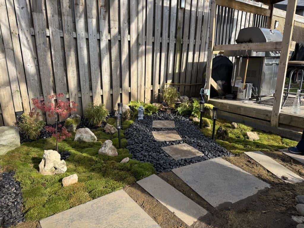 Small garden with stepping stones, black river rock and moss along a fence line